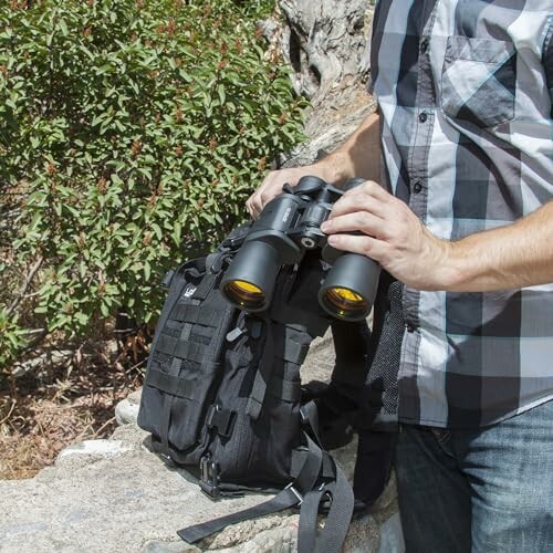 Person holding binoculars next to a backpack outdoors